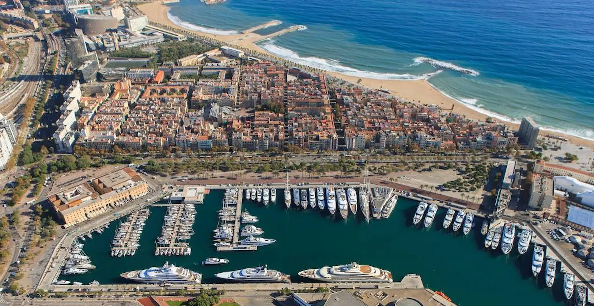 Aerial view of Marina Port Vell with numerous moored superyachts, Barcelona beach and Mediterranean Sea beyond.