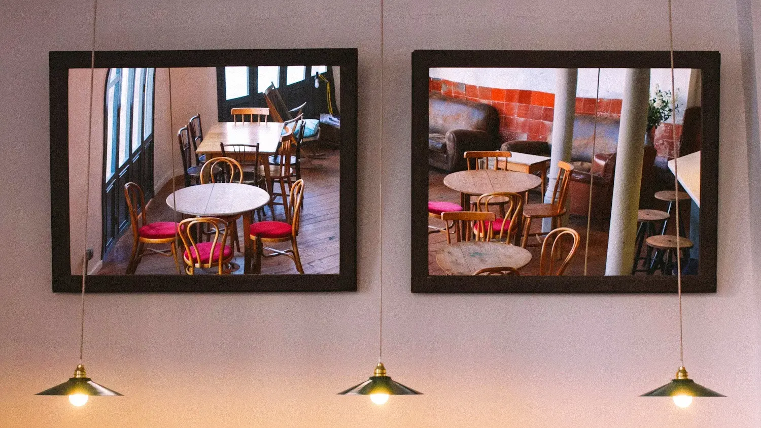 Two framed photographs of a modern caf&eacute; interior with round tables and wooden chairs hanging on a pink wall above brass pendant lights.
