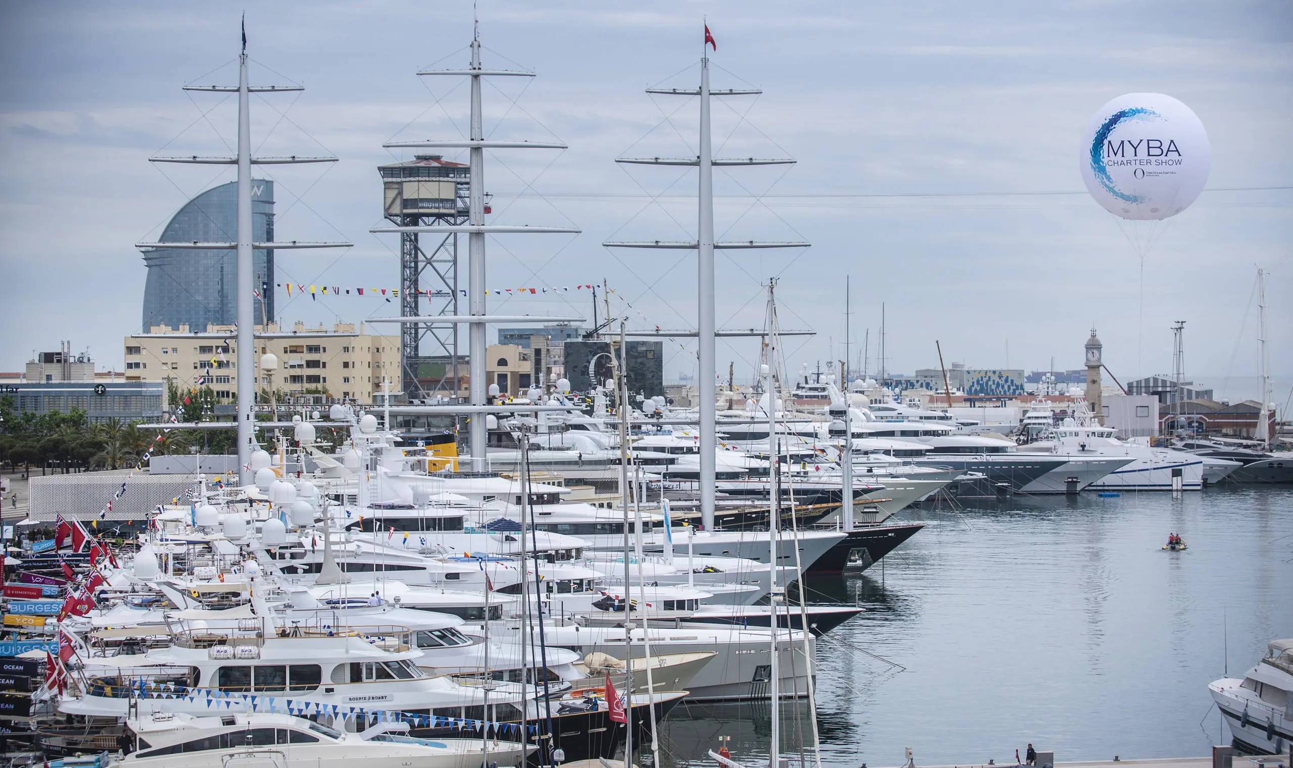Multiple superyachts moored at Marina Port Vell with tall masts and Barcelona waterfront buildings in the background.