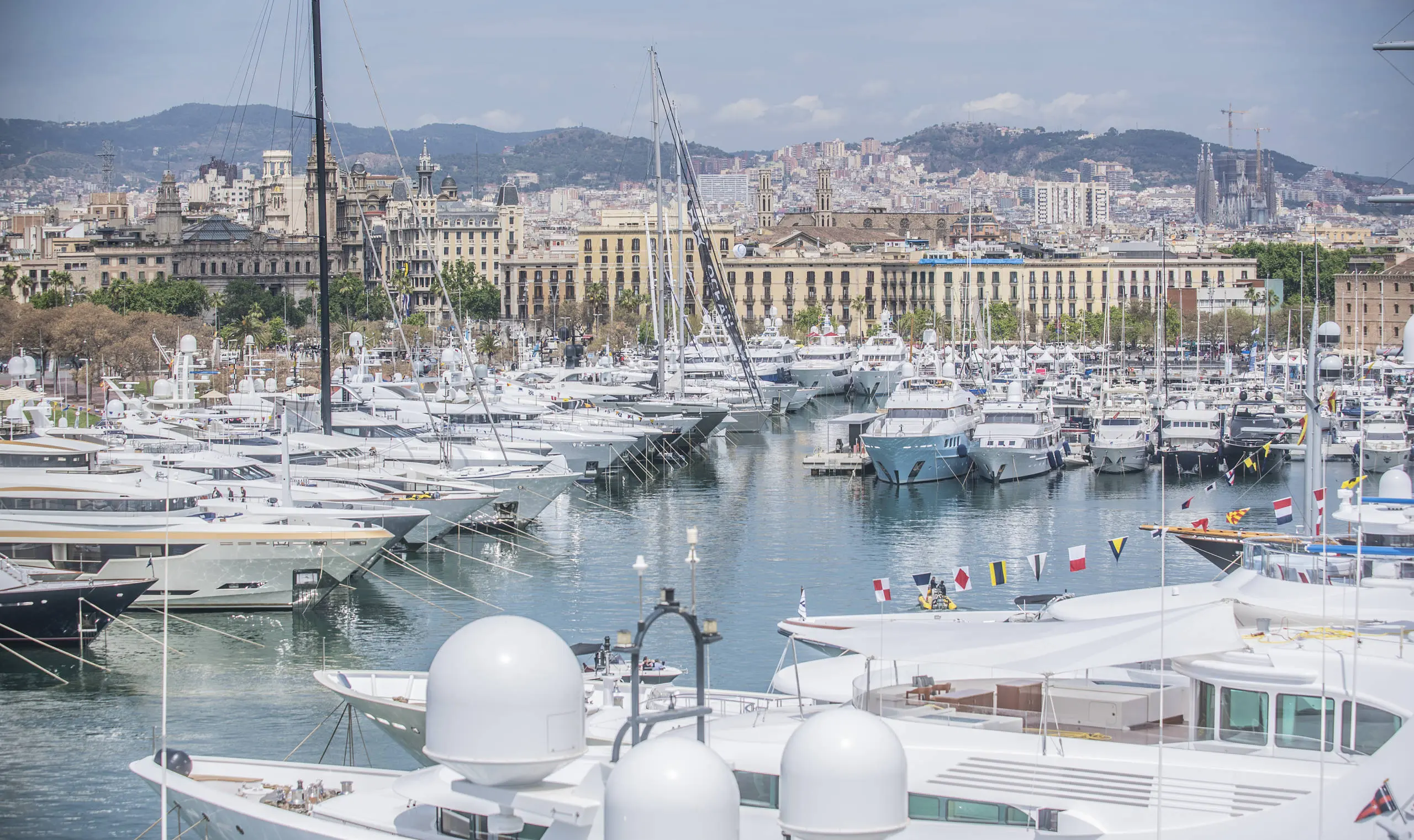Superyachts and sailboats moored in Barcelona marina with Gothic Quarter and city skyline in background.