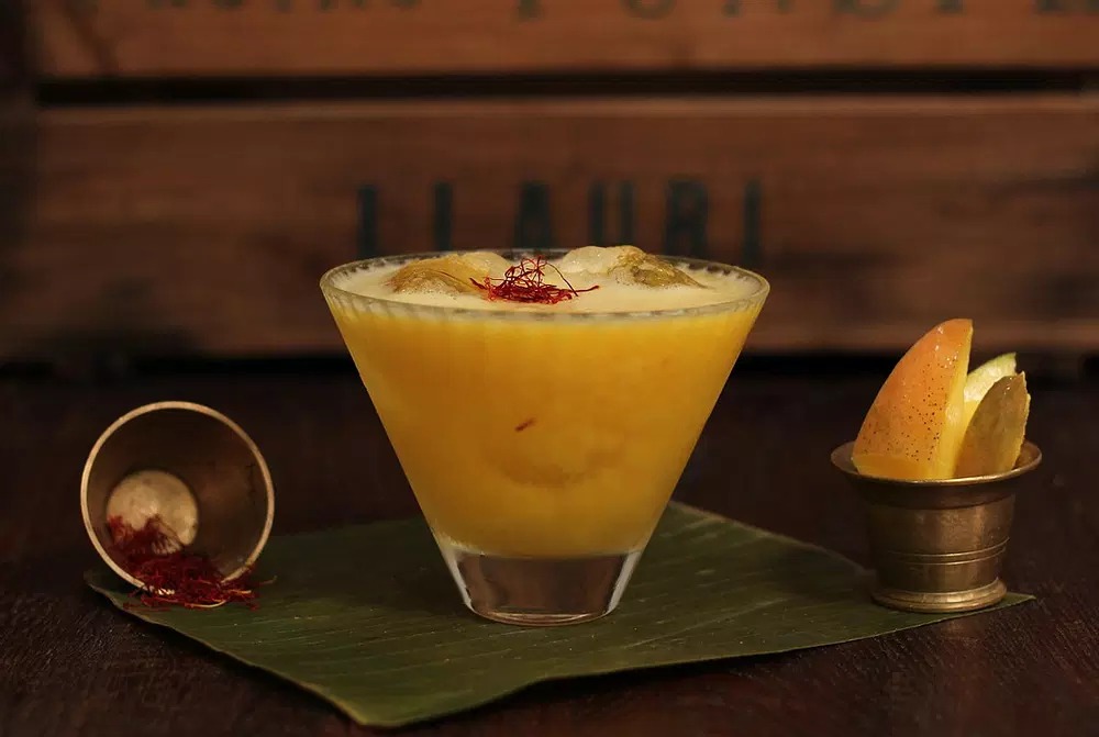 Yellow cocktail with spice garnish served with citrus fruit and decorative vessels on a green slate.
