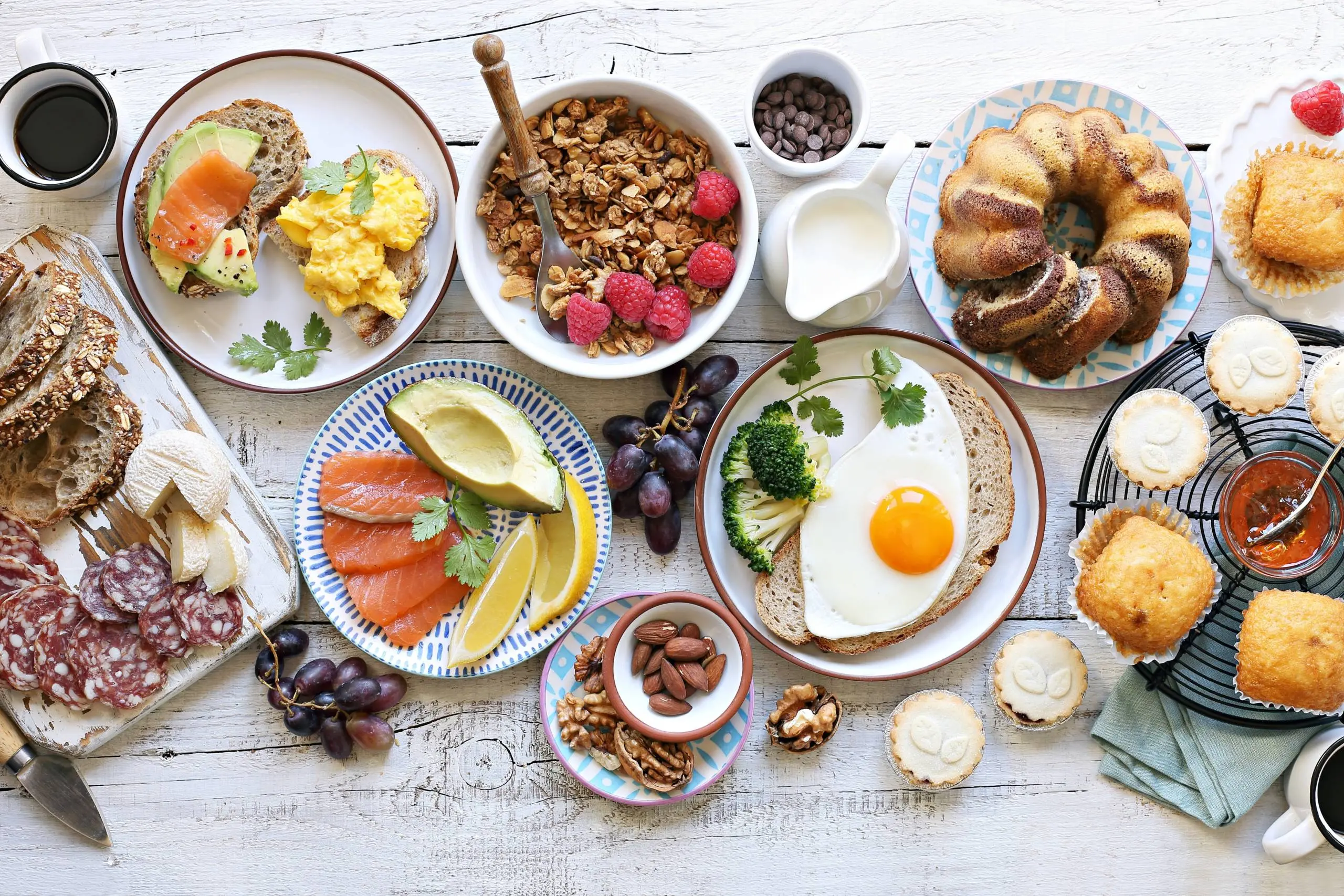 Overhead view of breakfast spread with toast, eggs, salmon, pastries, granola, nuts, fruit and cured meats.