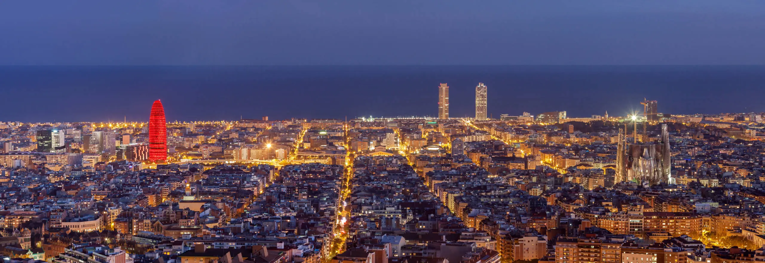 Barcelona cityscape at dusk with illuminated Torre Agbar, Sagrada Familia, and coastal horizon.
