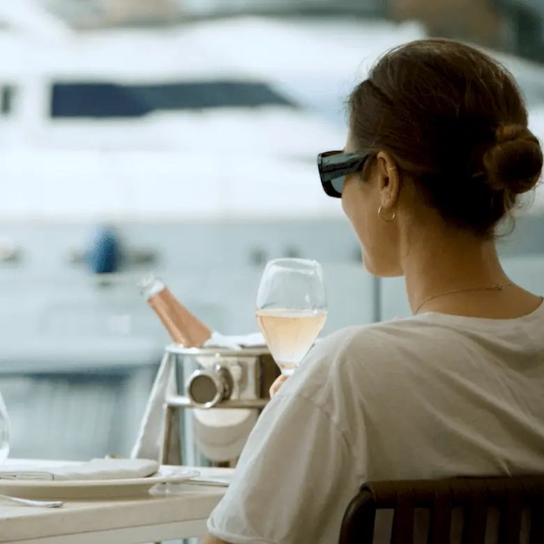 Woman in sunglasses sitting at waterfront table overlooking superyachts moored in marina.