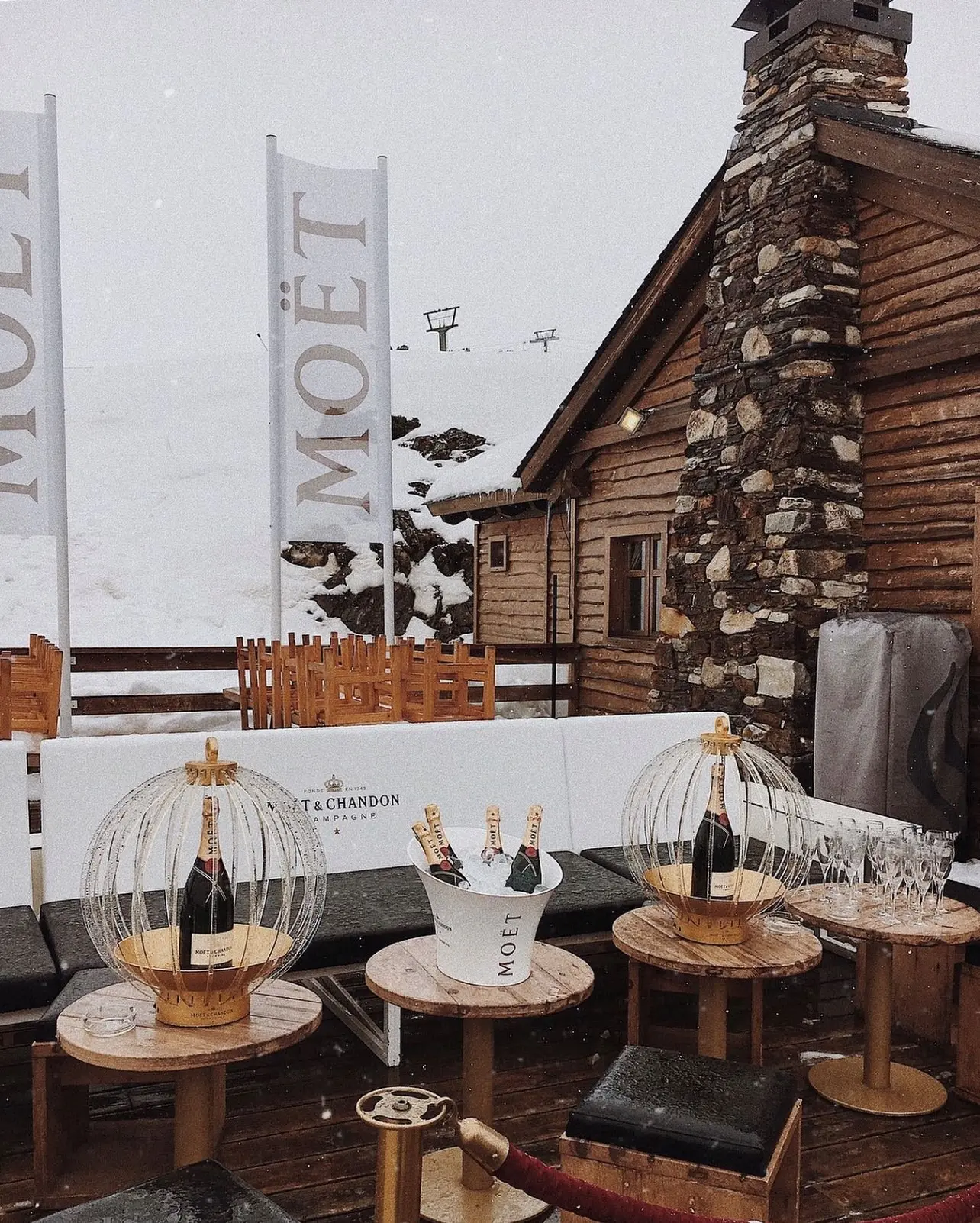 Champagne bottles displayed in decorative birdcage holders on a snowy mountain terrace with wooden cabins.