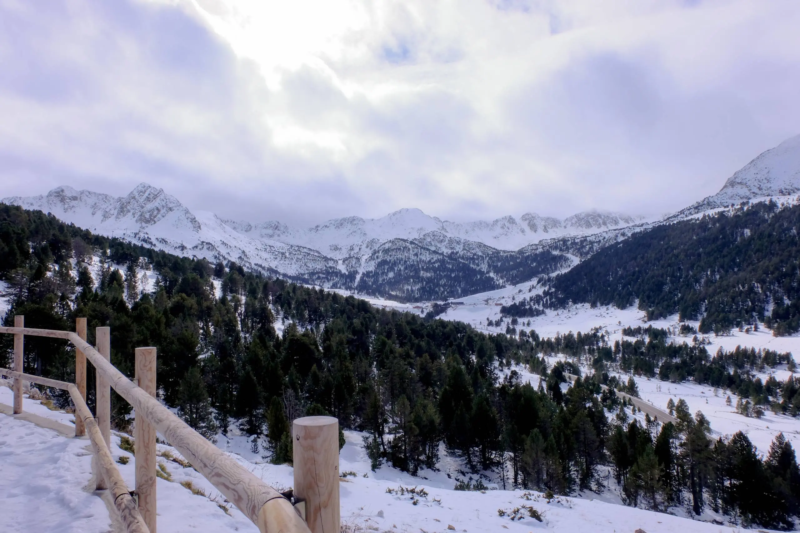 Snow-covered mountain valley with forested slopes and wooden fence railing in foreground.