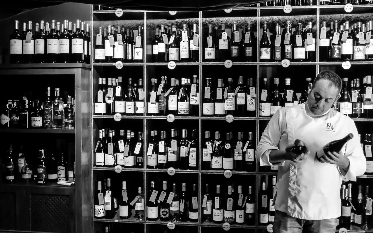 Man in white shirt checking wine bottle label in front of extensive wine cellar shelving.