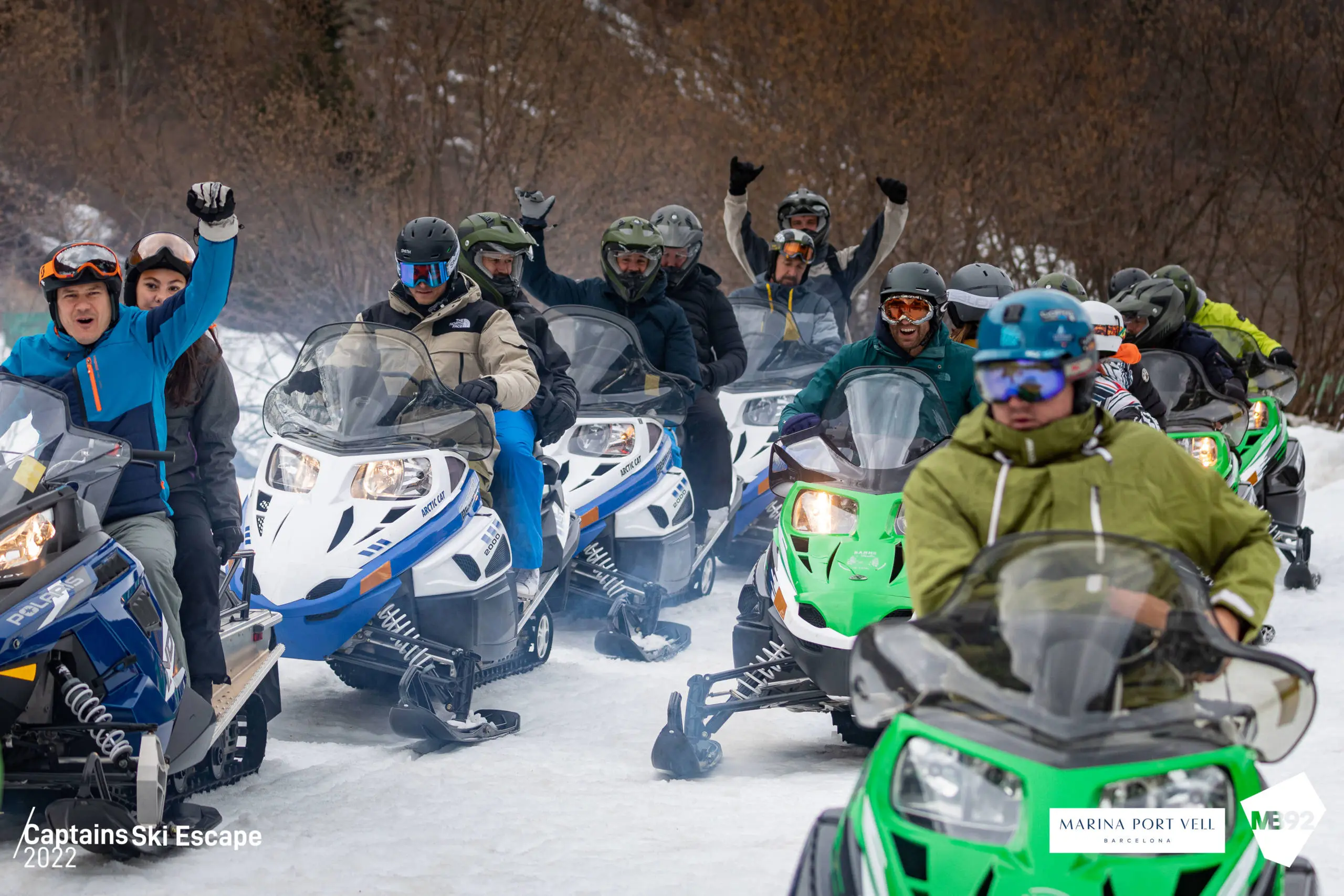Group of people on snowmobiles in snow-covered landscape with bare trees in background.