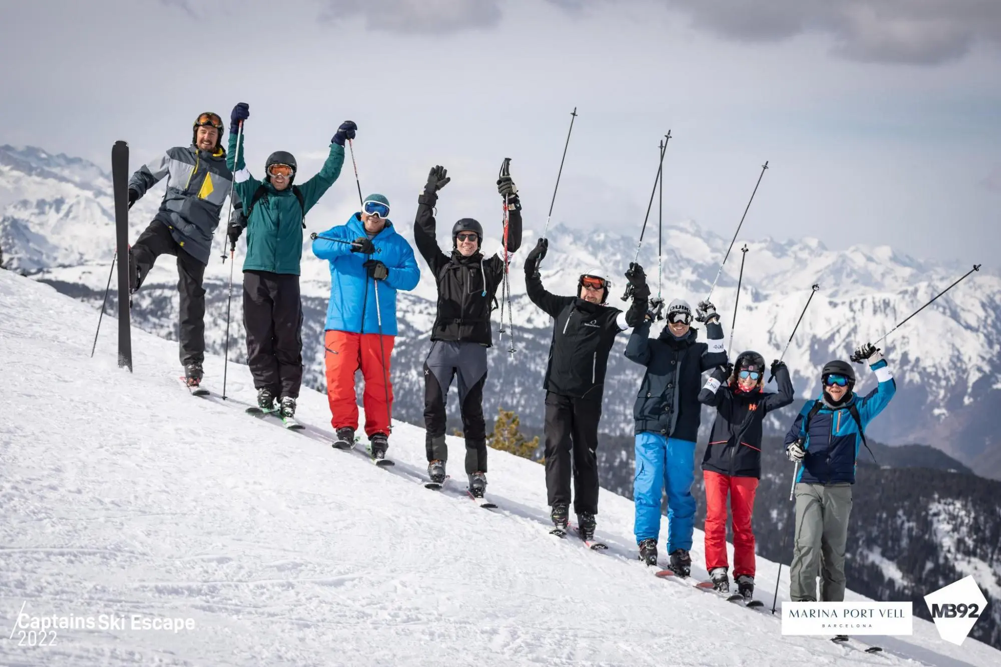 Group of skiers celebrating on a snowy mountain slope with snow-covered peaks in the background.