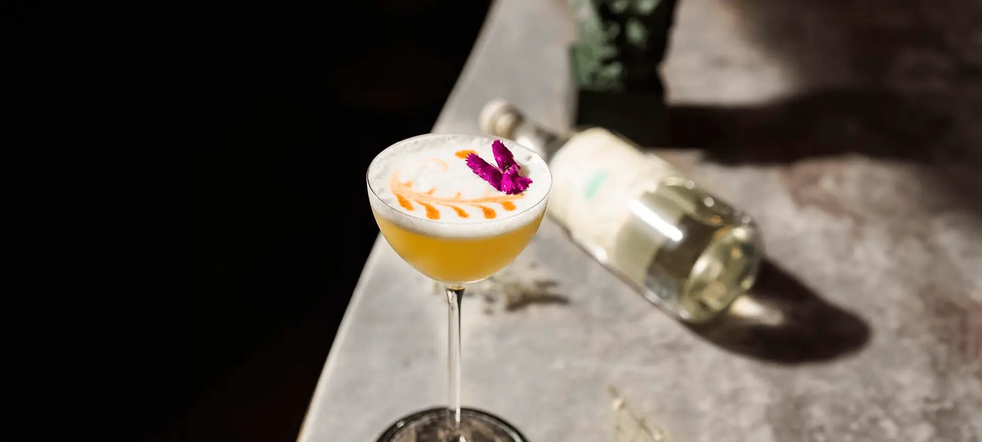 Cocktail with yellow liquid, foam top, and purple flower garnish beside a wine bottle.