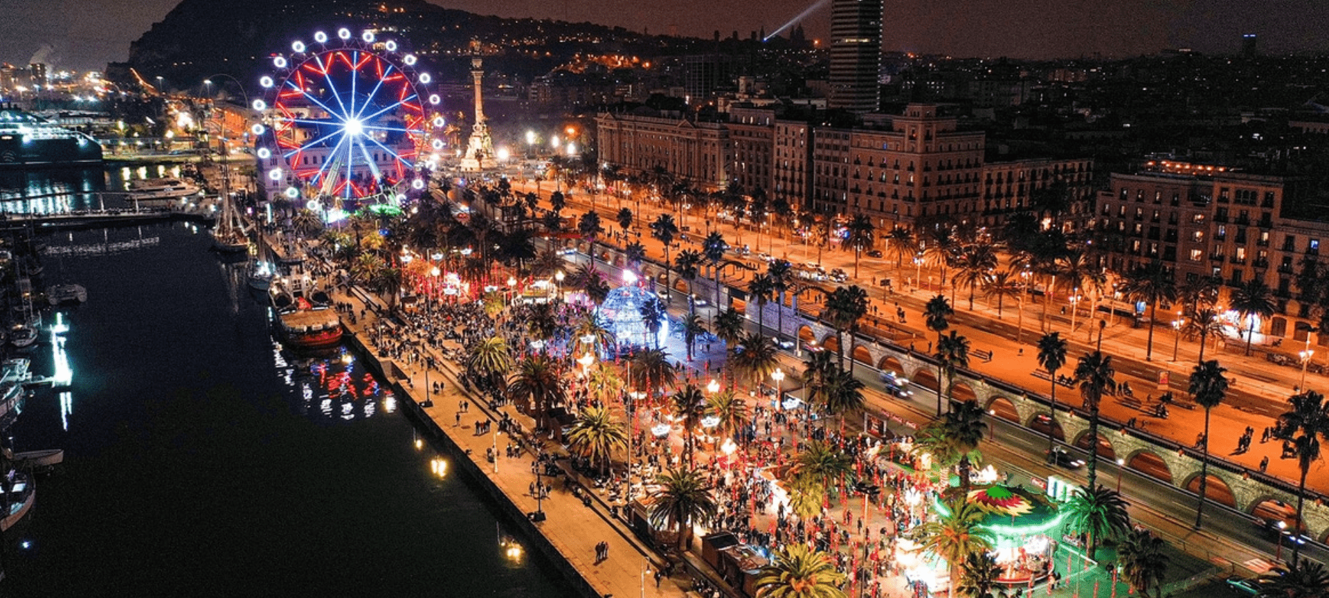Aerial drone view of the Christmas Fair (Fira de Nadal) at Barcelona’s Port Vell, featuring the illuminated Ferris wheel, festive stalls, and a lively holiday atmosphere by the sea